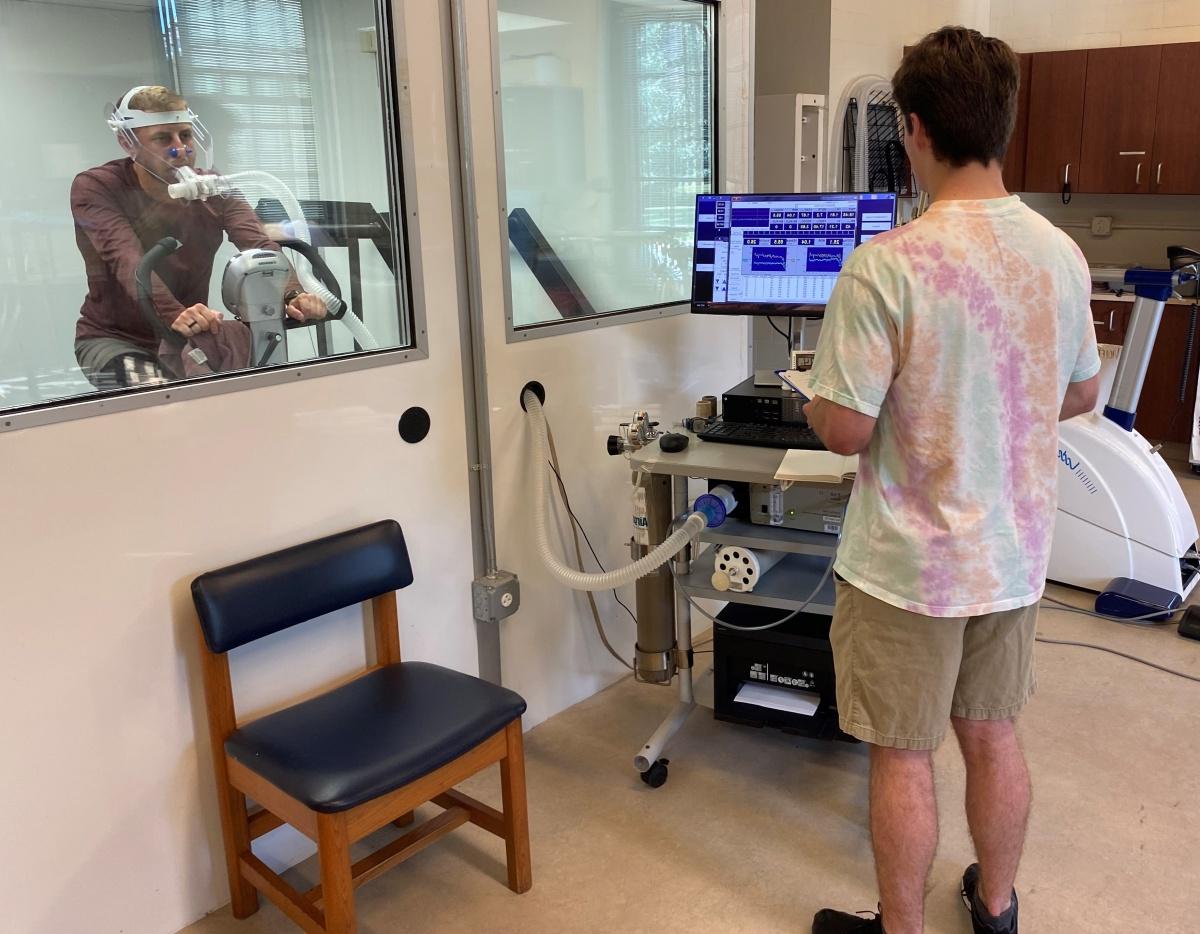 研究er looks at a computer while a subject rides an exercise bicycle behind glass