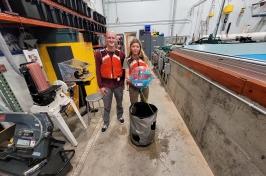 Two 主要研究 student researchers hold their winning ocean renewable energy device in front of a wave tank. They are wearing red life jackets.